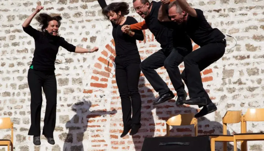 Vier Darsteller springen synchron in die Luft. Sie tragen schwarze Kleidung und Mikrofone. Ihre Mimik zeigt Freude und Energie, fast wie bei einem Playback-Auftritt.