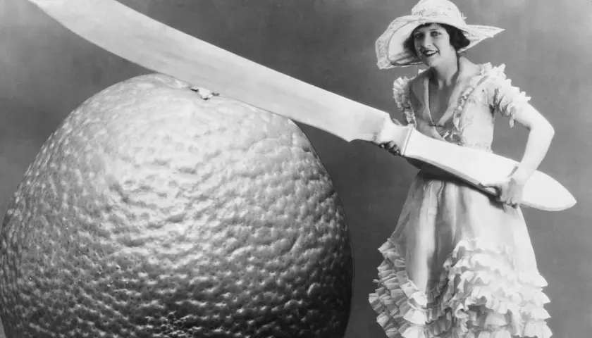 Eine Frau im Rüschenkleid hält ein riesiges Messer neben einer gigantischen Orange. Die Szene wirkt fast grotesk, eine humorvolle Darstellung von Größenverhältnissen.
