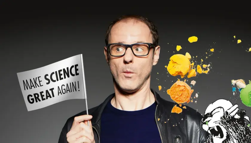 Vince Ebert mit Brille hält eine Fahne "Make Science Great Again!". Er blickt überrascht auf zerbrechende Macarons und eine Löwenzeichnung.