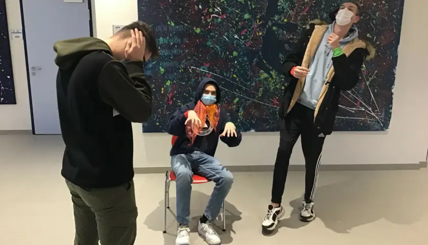 Drei Jungen mit Masken vor einem abstrakten Gemälde. Einer sitzt, die anderen stehen. Ein Moment wie ein Standbild im Theater.