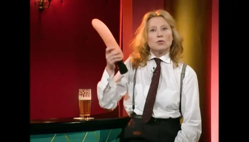 Eine Frau in einem weißen Hemd mit Krawatte und Hosenträgern hält einen rosafarbenen Gegenstand hoch. Was ist Sketch? Ein Glas Bier steht daneben.