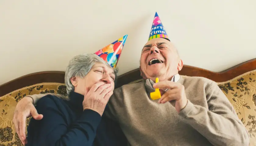 Ein älteres Paar mit Partyhüten lacht herzhaft. Sie zeigen, wie man jemanden zum Lachen bringt: mit Freude und Humor.
