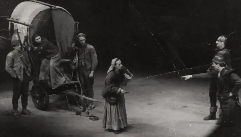 Szene aus einem **Brecht Theater**: Eine Frau mit Kopftuch und Korb steht vor einem Wagen. Zwei Männer in Rüstung zeigen in eine Richtung.