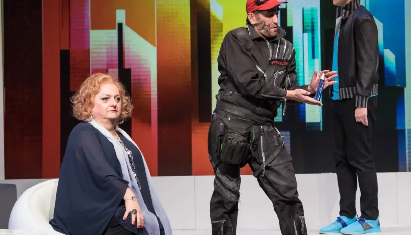 Ein Mann mit roter Kappe und Werkzeuggürtel reicht einer sitzenden Frau eine Sprühflasche. Ein Standbild-Beispiel einer Theateraufführung.