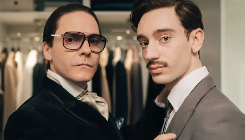 Zwei Männer in eleganter Kleidung, einer mit Brille und Krawatte, der andere mit Schnurrbart, scheinen auf dem Weg zu sein, Karl Lagerfeld zu werden.
