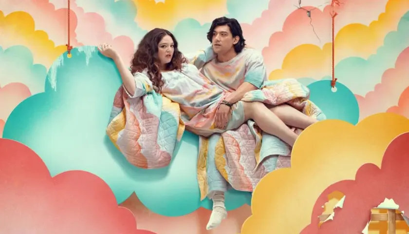 Ein Paar sitzt auf einer Wolke, eingehüllt in eine bunte Decke. Die besetzung von Too Much scheint in einer surrealen Traumwelt zu schweben.