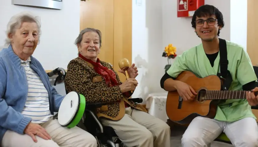 Zwei ältere Damen und ein junger Mann musizieren gemeinsam. Die Damen spielen Trommel und Maracas, der Mann spielt Gitarre. Ein fröhlicher Moment, wie im **Coco Film**.