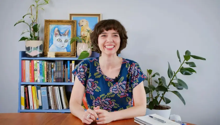 Eine Frau mit einem breiten Lächeln sitzt vor einem Bücherregal mit Katzen- und Hundebildern. Sie wirkt aufgeregt und lustig, bereit für ein Gespräch.