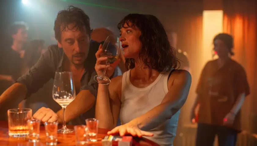 Ein Mann und eine Frau sitzen an einer Bar, sie trinkt aus einem Weinglas. Die Szene erinnert an den Film "der rausch film", mit gedämpftem Licht und einer intimen Atmosphäre.