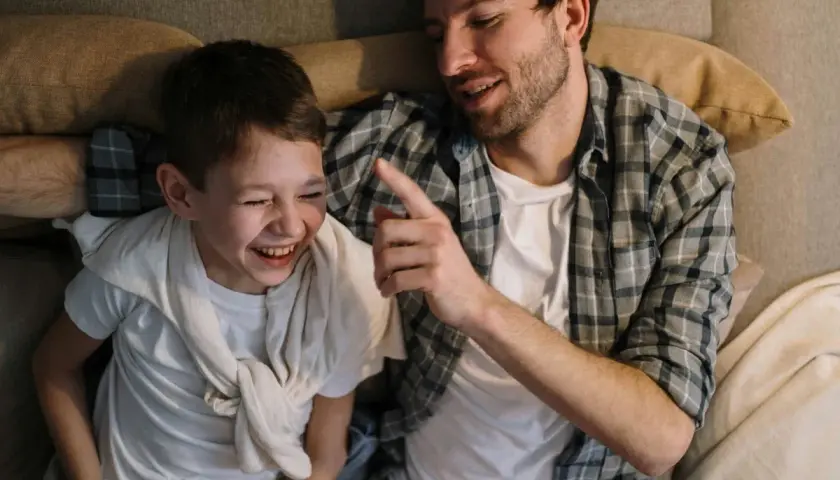 Ein Vater erzählt seinem Sohn einen Knock-Knock-Witz. Der Junge lacht herzhaft, während der Vater ihm mit einem Finger deutet.