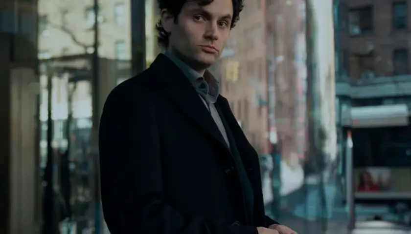 Joe Goldberg aus der serie "You" steht nachdenklich in Schwarz gekleidet vor einem modernen Gebäude.
