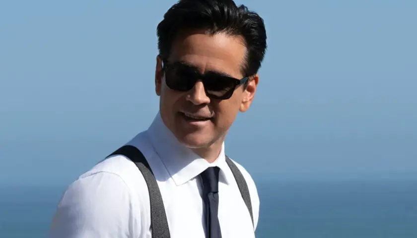 John Sugar, mit Sonnenbrille und Hosenträgern, lächelt vor dem blauen Himmel und Meer.