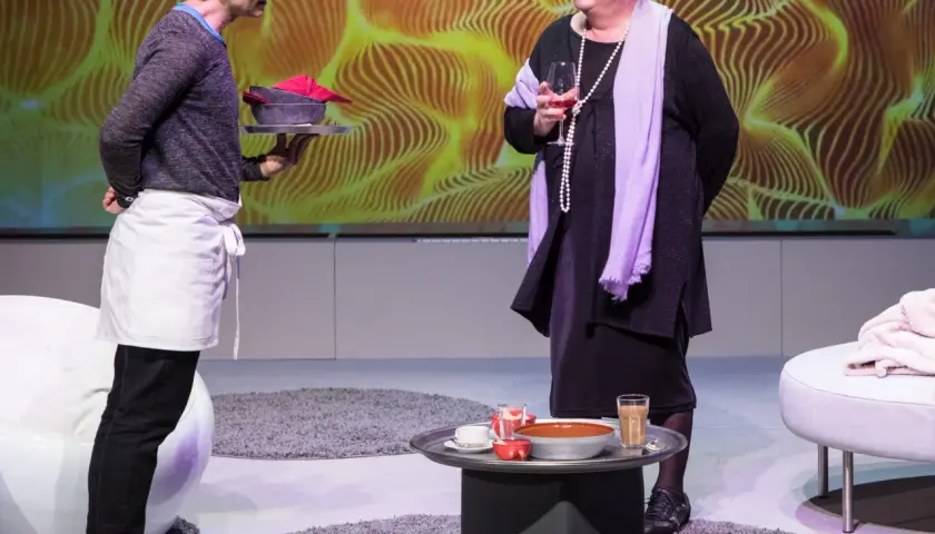 Zwei Personen stehen vor einer gelben, wellenförmigen Kulisse. Eine Person mit Schürze trägt ein Tablett mit Schalen, die andere hält ein Glas Wein. Ein scheiter heiter Moment.