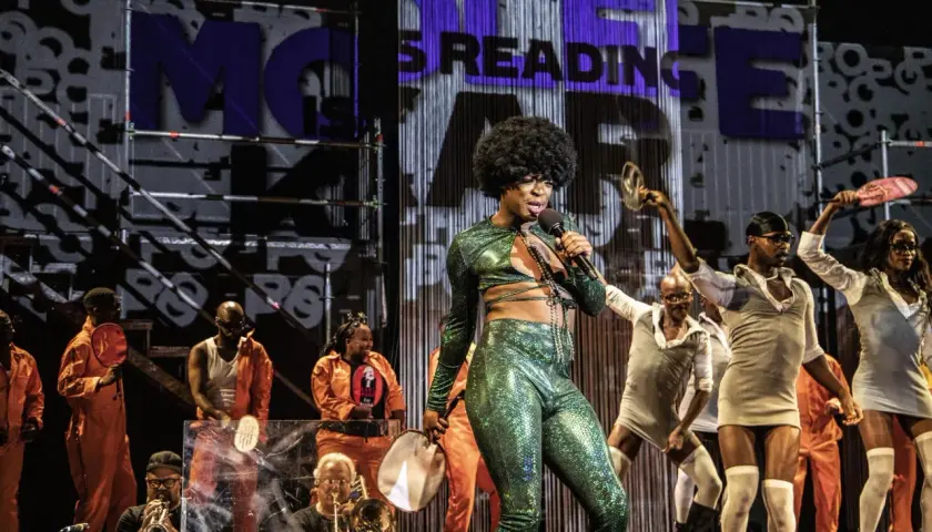 Künstler mit Afro-Perücke singt in glitzerndem Outfit. Im Hintergrund tanzen Menschen, einige in orangefarbenen Overalls. Was ist Monolog? Eine Performance, die fesselt.