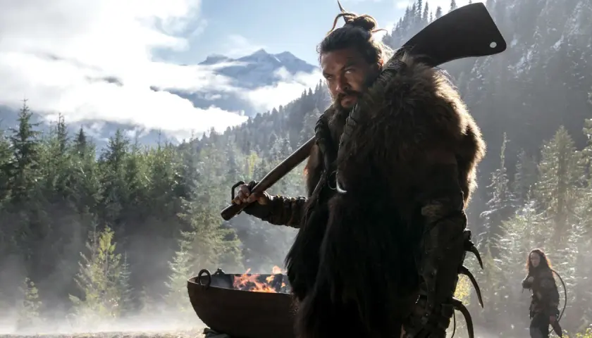 Mann mit Fellmantel und Axt blickt ernst in die Kamera, im Hintergrund Berge und Wald. Ein Feuer brennt in einem Kessel. Sieh diese Serie!