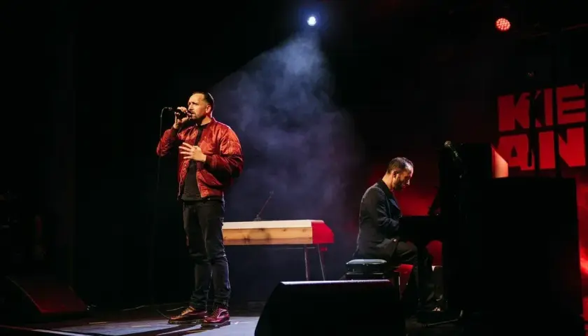 Ein Sänger mit Mikrofon und ein Pianist auf der Bühne. Die Beleuchtung schafft eine dramatische Atmosphäre, die gut zu spontanen **Stegreifrede Themen** passt.