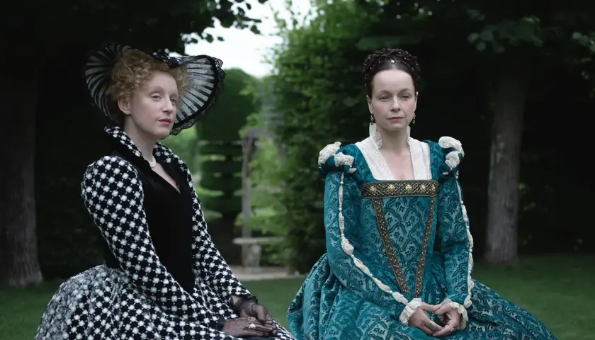 Zwei Frauen in historischen Gewändern, eine in Schwarz-Weiß, die andere in Türkis, sitzen im Garten. Die eine könnte die Schlangen-Königin sein, die andere ihre Rivalin.