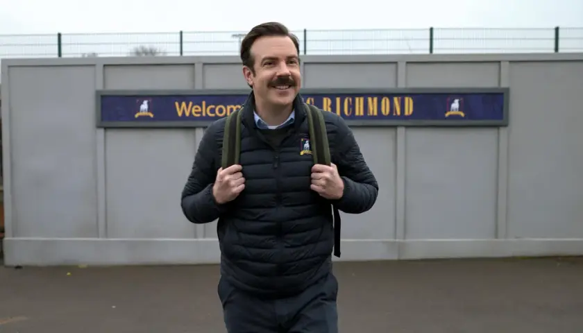 Ted Lasso, mit seinem charakteristischen Schnurrbart, trägt einen Rucksack vor dem Schild "Welcome to Richmond".