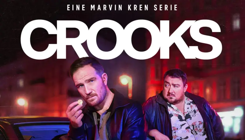 Zwei Männer, Teil der "Crooks"-Besetzung, stehen im nächtlichen Stadtlicht. Einer hält eine Münze, der andere blickt nachdenklich.