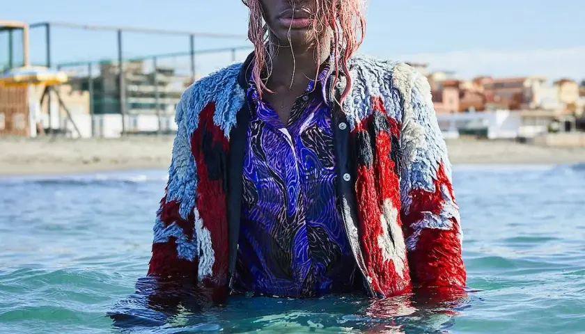 Ein Model mit pinken Haaren steht im Meer, trägt eine bunte Jacke und ein blaues Hemd. Die Wellen umspielen sie, als ob sie sagen: "I may destroy you.