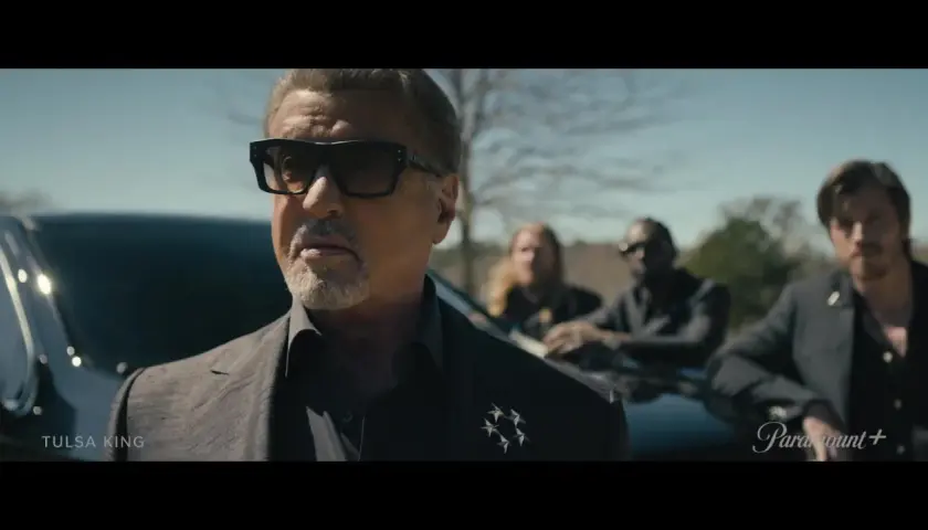 Sylvester Stallone als Tulsa King, mit Sonnenbrille und Anzug, steht vor einem Auto, begleitet von drei Männern.