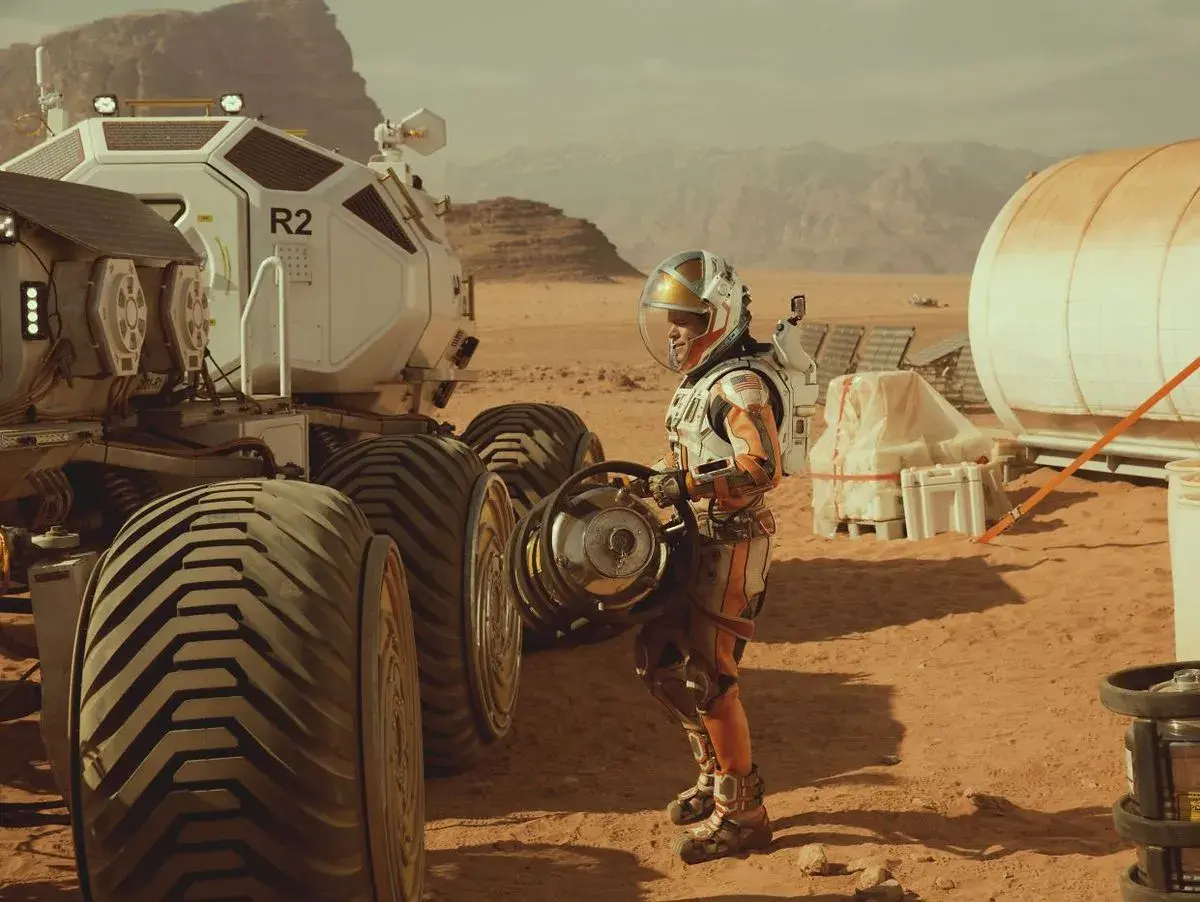 Ein Astronaut in einem orangefarbenen Raumanzug steht neben einem Mars-Rover. Szene aus dem Marsianer Film.