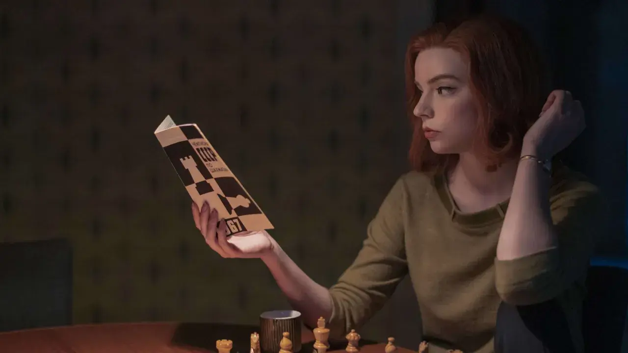 Beth Harmon studiert eine Schachpartie, inspiriert vom Damengambit. Schachfiguren und eine Tasse stehen auf dem Tisch.