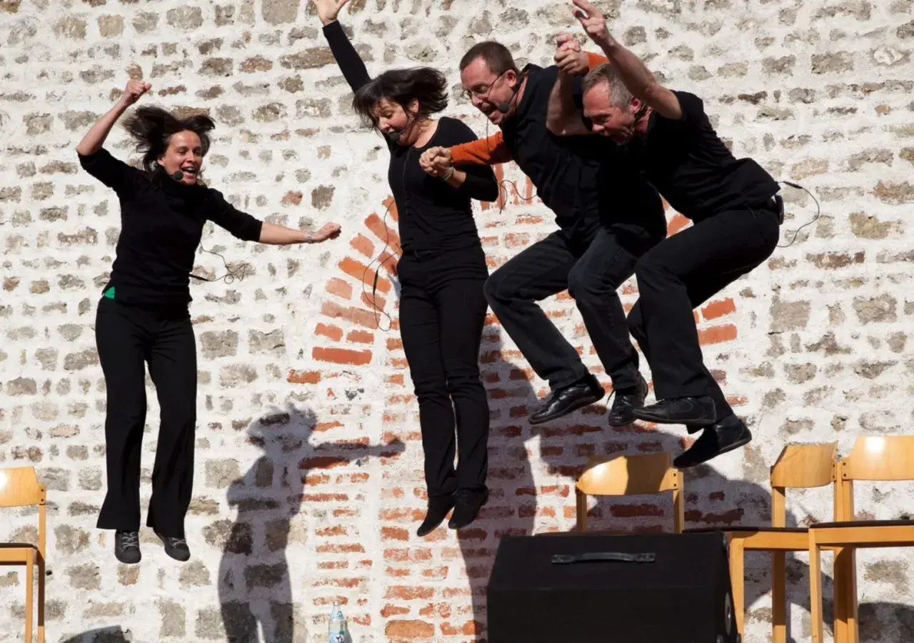 Vier Darsteller springen synchron in die Luft. Sie tragen schwarze Kleidung und Mikrofone. Ihre Mimik zeigt Freude und Energie, fast wie bei einem Playback-Auftritt.