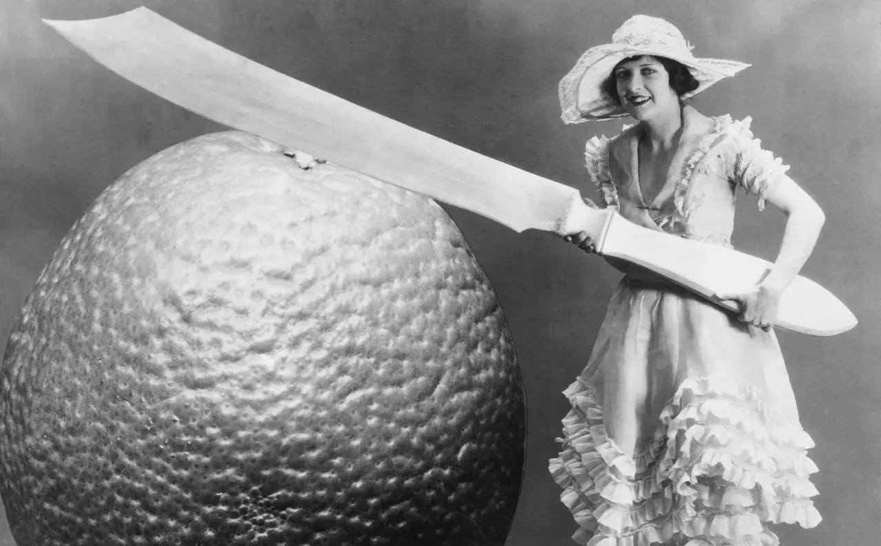 Eine Frau im Rüschenkleid hält ein riesiges Messer neben einer gigantischen Orange. Die Szene wirkt fast grotesk, eine humorvolle Darstellung von Größenverhältnissen.