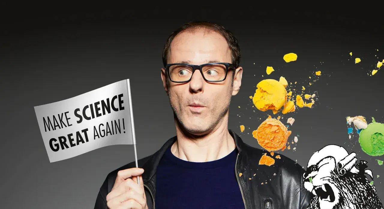 Vince Ebert mit Brille hält eine Fahne "Make Science Great Again!". Er blickt überrascht auf zerbrechende Macarons und eine Löwenzeichnung.
