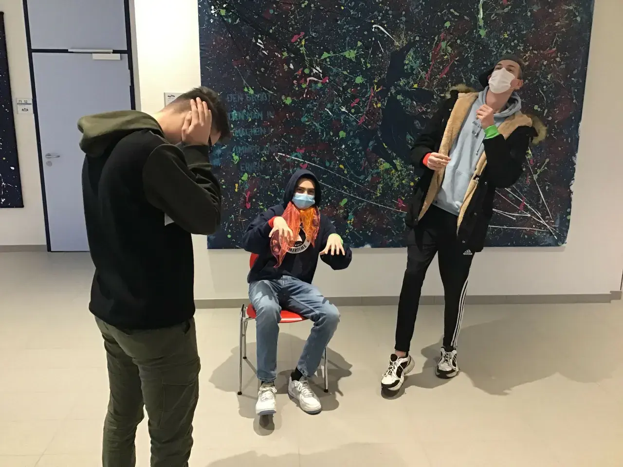 Drei Jungen mit Masken vor einem abstrakten Gemälde. Einer sitzt, die anderen stehen. Ein Moment wie ein Standbild im Theater.