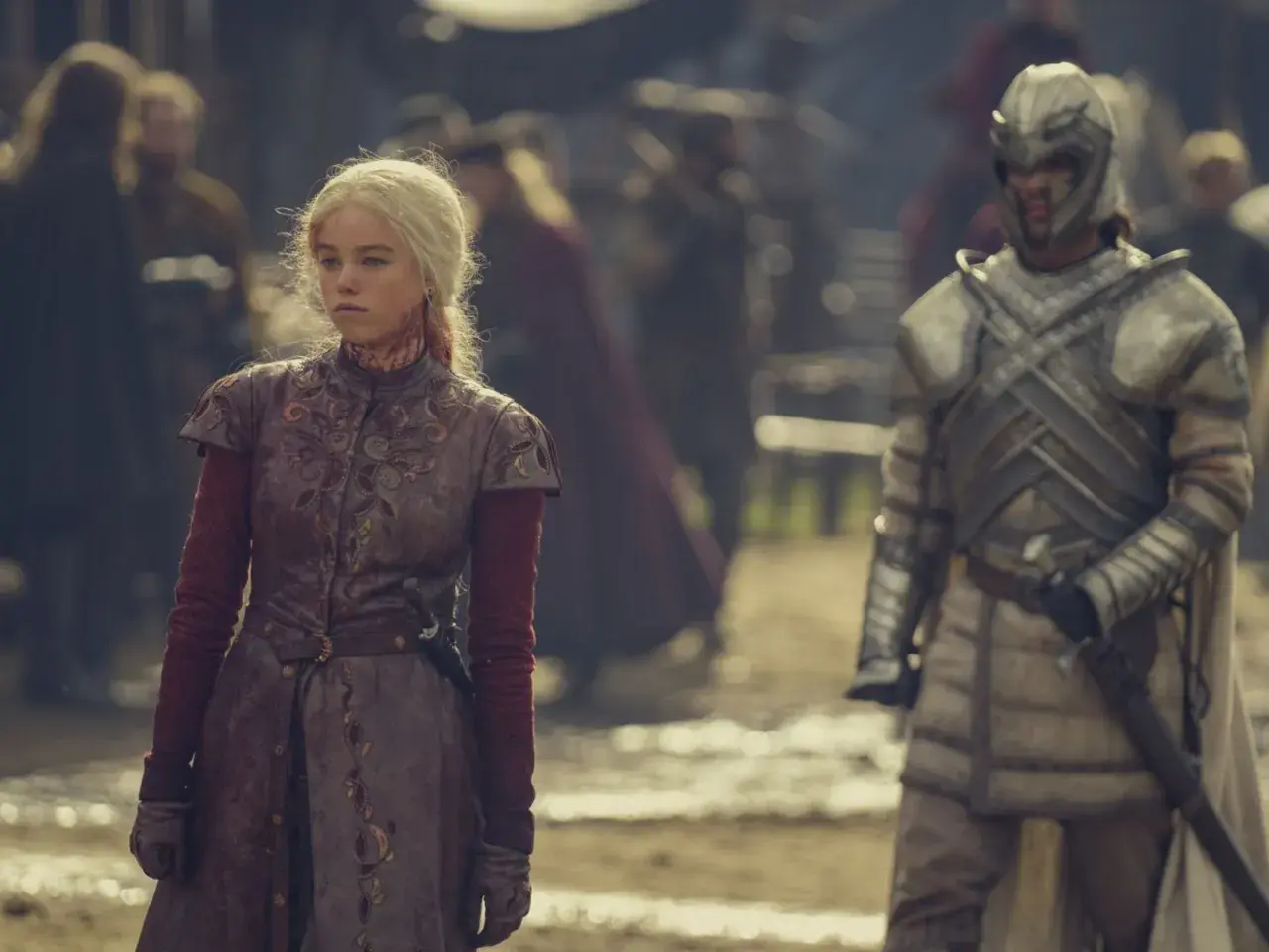 Rhaenyra Targaryen, blutverschmiert, blickt nachdenklich, während ein Ritter in voller Rüstung im Hintergrund der Folgen von House of the Dragon steht.