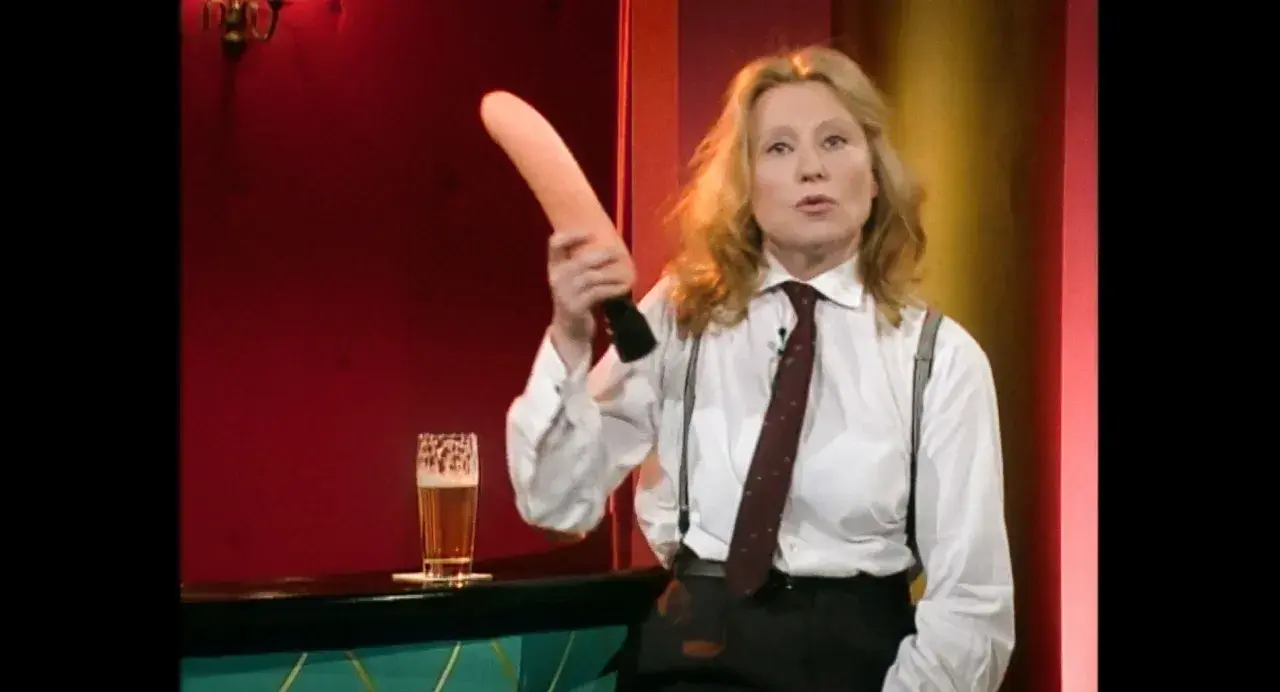 Eine Frau in einem weißen Hemd mit Krawatte und Hosenträgern hält einen rosafarbenen Gegenstand hoch. Was ist Sketch? Ein Glas Bier steht daneben.