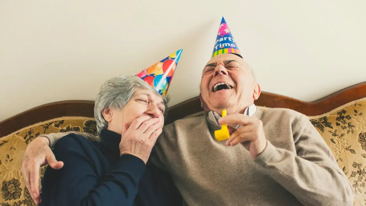 Ein älteres Paar mit Partyhüten lacht herzhaft. Sie zeigen, wie man jemanden zum Lachen bringt: mit Freude und Humor.