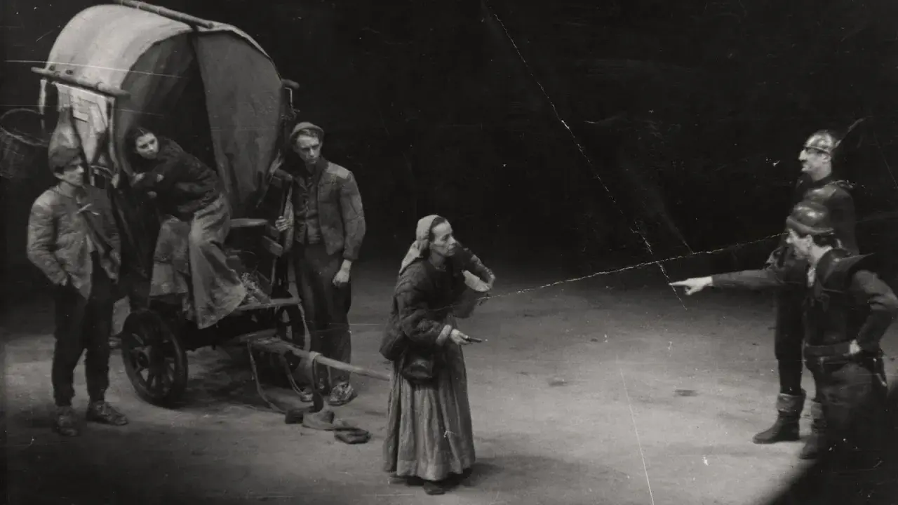 Szene aus einem **Brecht Theater**: Eine Frau mit Kopftuch und Korb steht vor einem Wagen. Zwei Männer in Rüstung zeigen in eine Richtung.