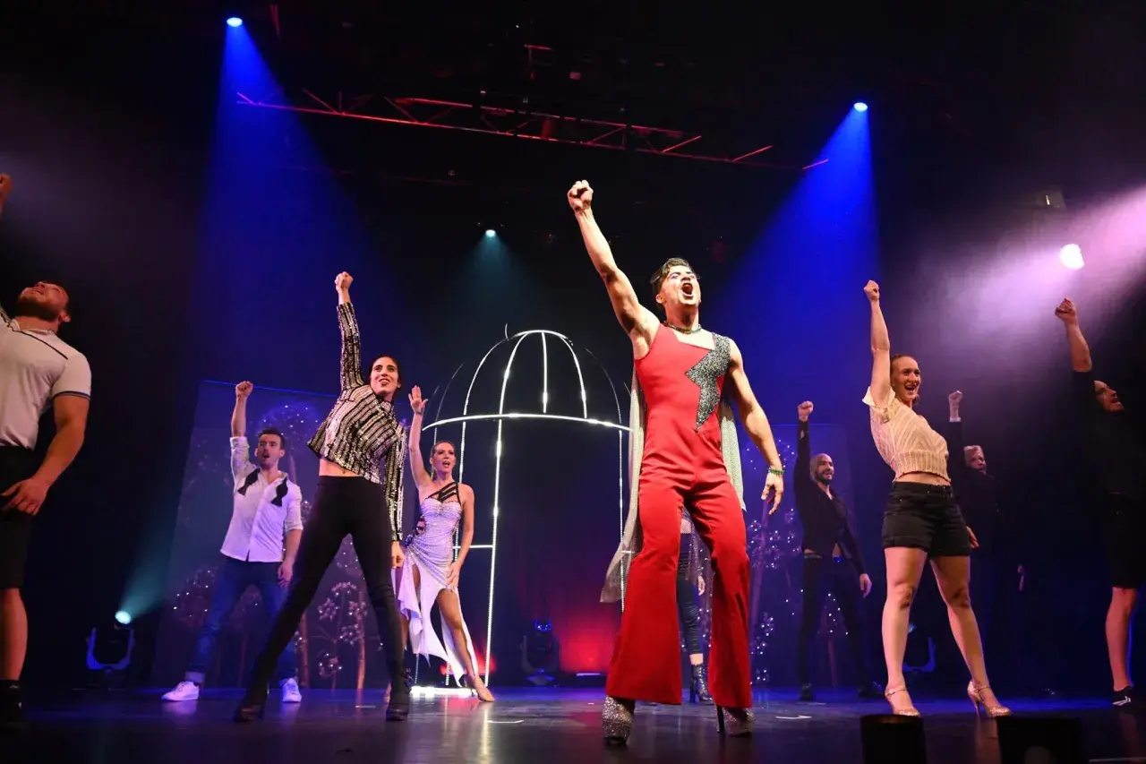 Künstler jubeln auf der Bühne bei einer **playback show**. Ein Mann in Rot dominiert das Bild.