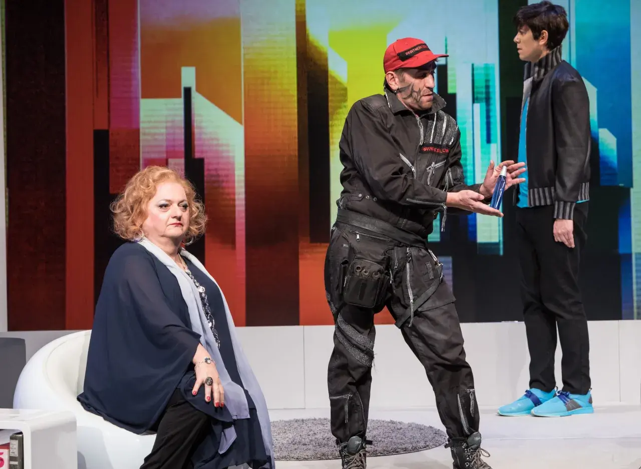Ein Mann mit roter Kappe und Werkzeuggürtel reicht einer sitzenden Frau eine Sprühflasche. Ein Standbild-Beispiel einer Theateraufführung.