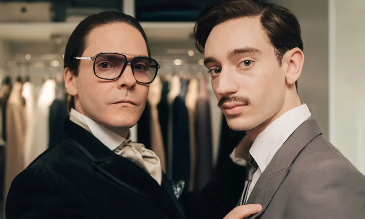 Zwei Männer in eleganter Kleidung, einer mit Brille und Krawatte, der andere mit Schnurrbart, scheinen auf dem Weg zu sein, Karl Lagerfeld zu werden.