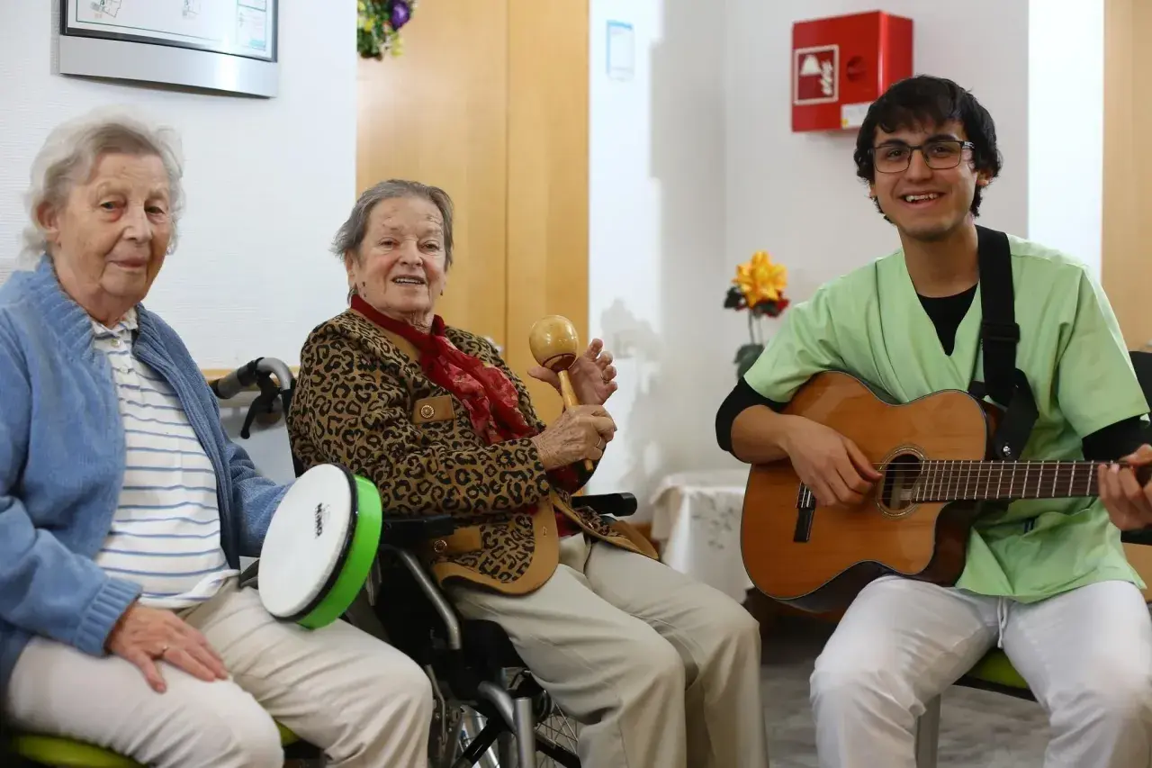 Zwei ältere Damen und ein junger Mann musizieren gemeinsam. Die Damen spielen Trommel und Maracas, der Mann spielt Gitarre. Ein fröhlicher Moment, wie im **Coco Film**.