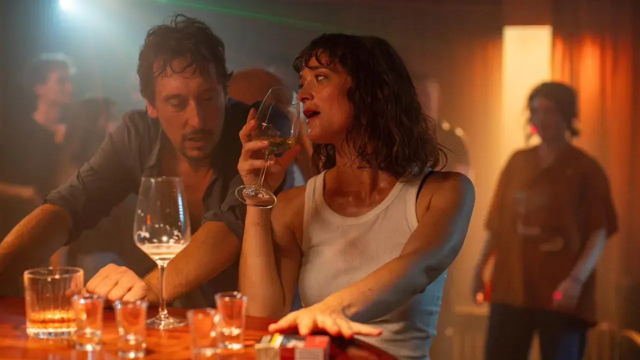 Ein Mann und eine Frau sitzen an einer Bar, sie trinkt aus einem Weinglas. Die Szene erinnert an den Film "der rausch film", mit gedämpftem Licht und einer intimen Atmosphäre.