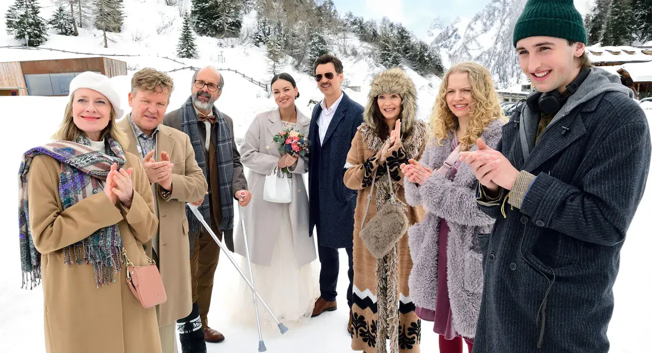 Die besetzung von der spitzname feiert eine Hochzeit im Schnee. Eine Braut mit Blumenstrauß, ein Bräutigam mit Schnurrbart und Gäste in Wintermänteln.