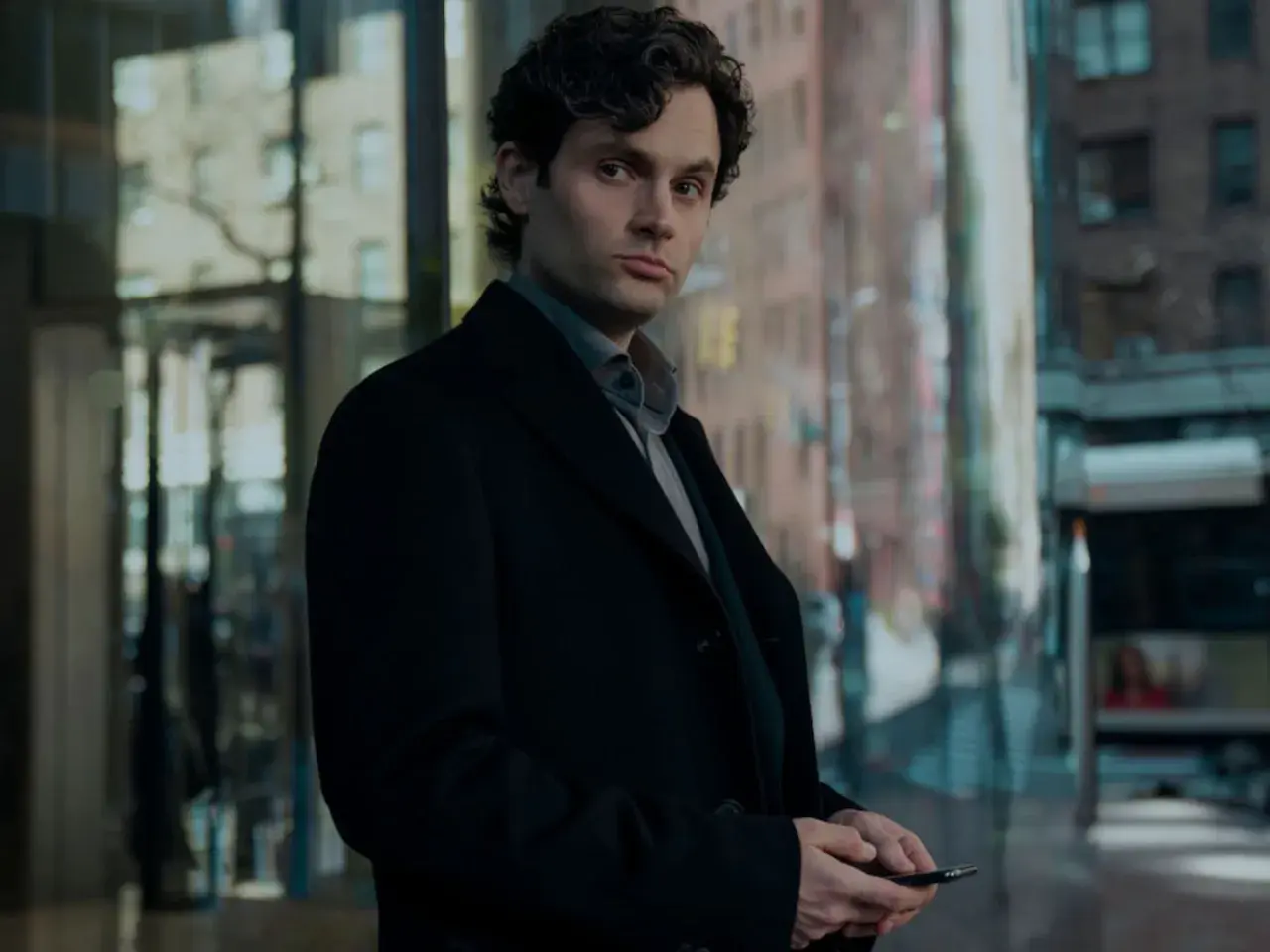 Joe Goldberg aus der serie "You" steht nachdenklich in Schwarz gekleidet vor einem modernen Gebäude.