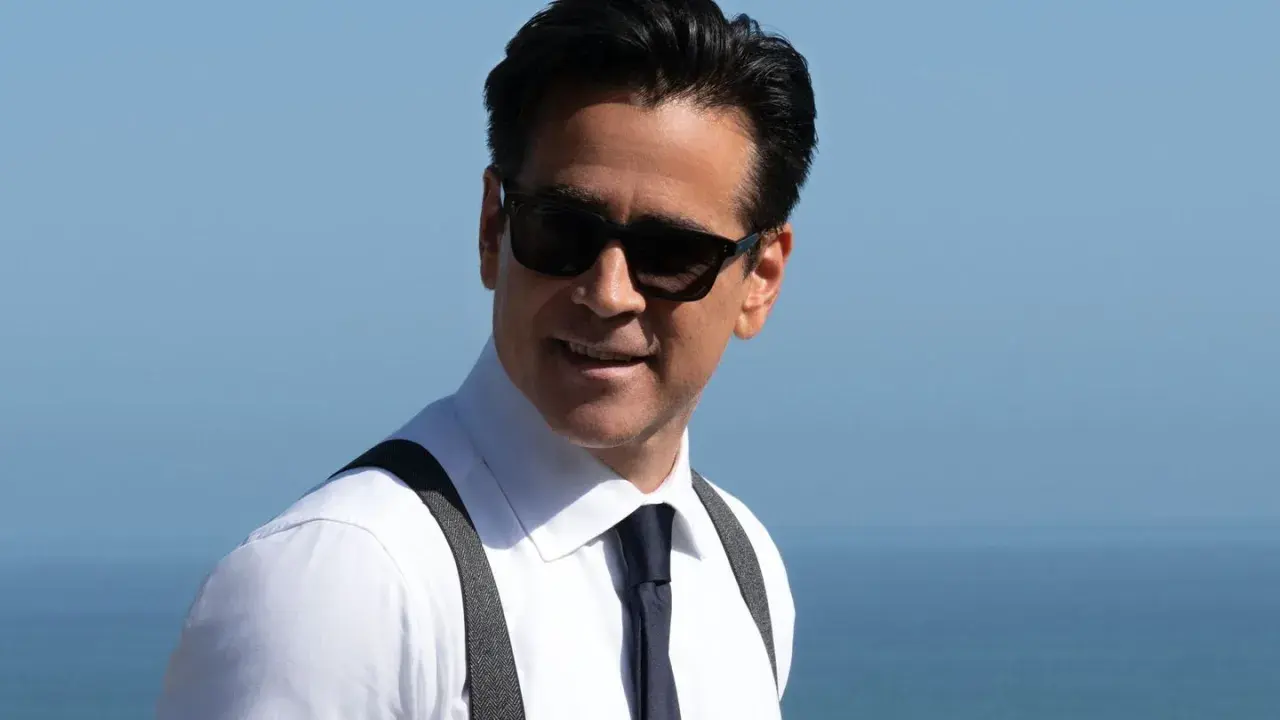 John Sugar, mit Sonnenbrille und Hosenträgern, lächelt vor dem blauen Himmel und Meer.