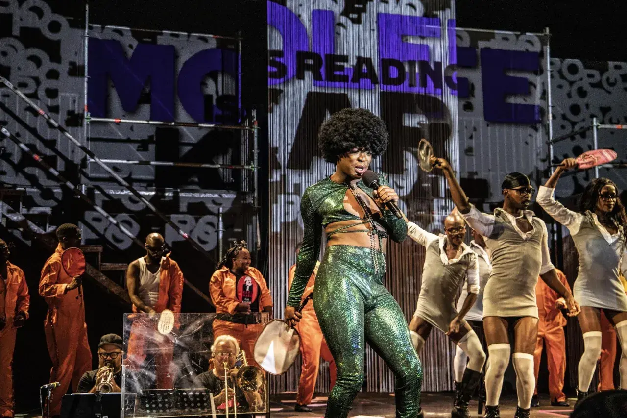 Künstler mit Afro-Perücke singt in glitzerndem Outfit. Im Hintergrund tanzen Menschen, einige in orangefarbenen Overalls. Was ist Monolog? Eine Performance, die fesselt.