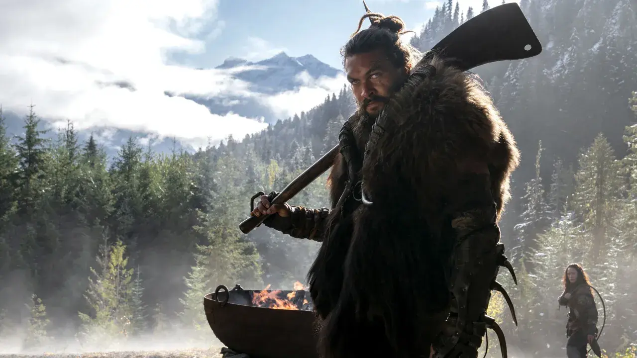 Mann mit Fellmantel und Axt blickt ernst in die Kamera, im Hintergrund Berge und Wald. Ein Feuer brennt in einem Kessel. Sieh diese Serie!