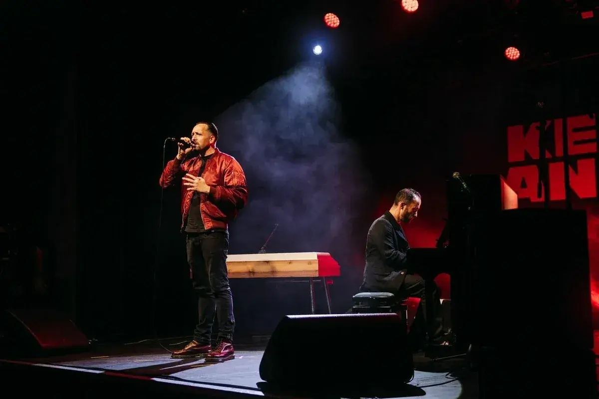 Ein Sänger mit Mikrofon und ein Pianist auf der Bühne. Die Beleuchtung schafft eine dramatische Atmosphäre, die gut zu spontanen **Stegreifrede Themen** passt.