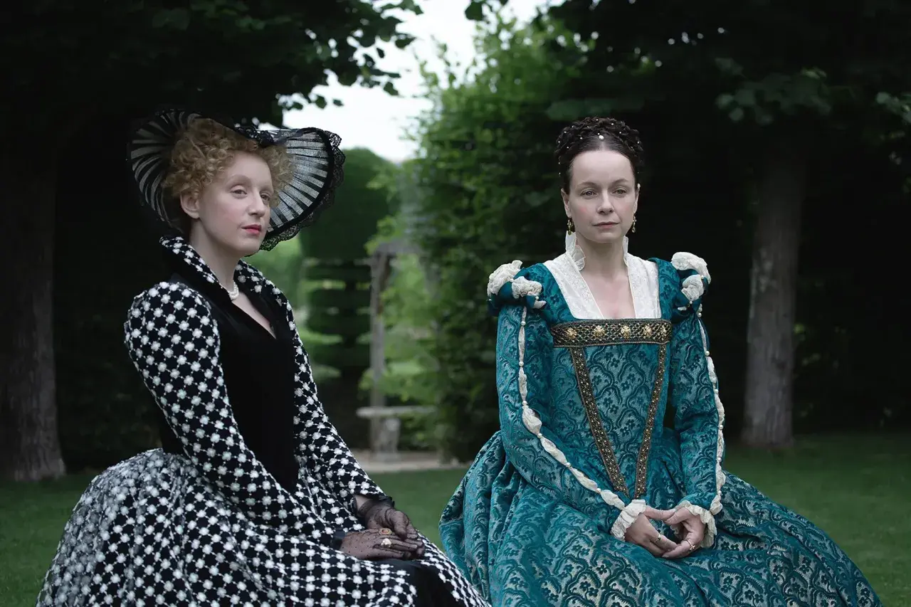 Zwei Frauen in historischen Gewändern, eine in Schwarz-Weiß, die andere in Türkis, sitzen im Garten. Die eine könnte die Schlangen-Königin sein, die andere ihre Rivalin.