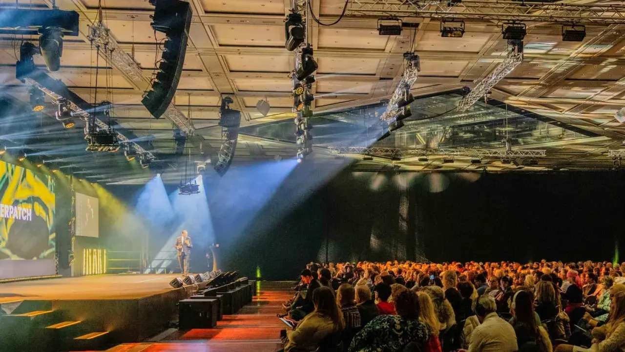 Bühne mit Redner, Publikum und Lichtshow. Die Atmosphäre erinnert an eine spontane Improvisation, Musik und ein inspirierendes Event.