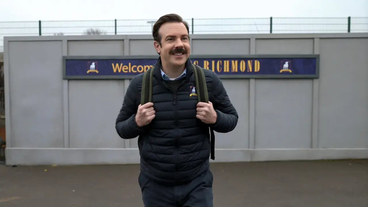 Ted Lasso, mit seinem charakteristischen Schnurrbart, trägt einen Rucksack vor dem Schild "Welcome to Richmond".
