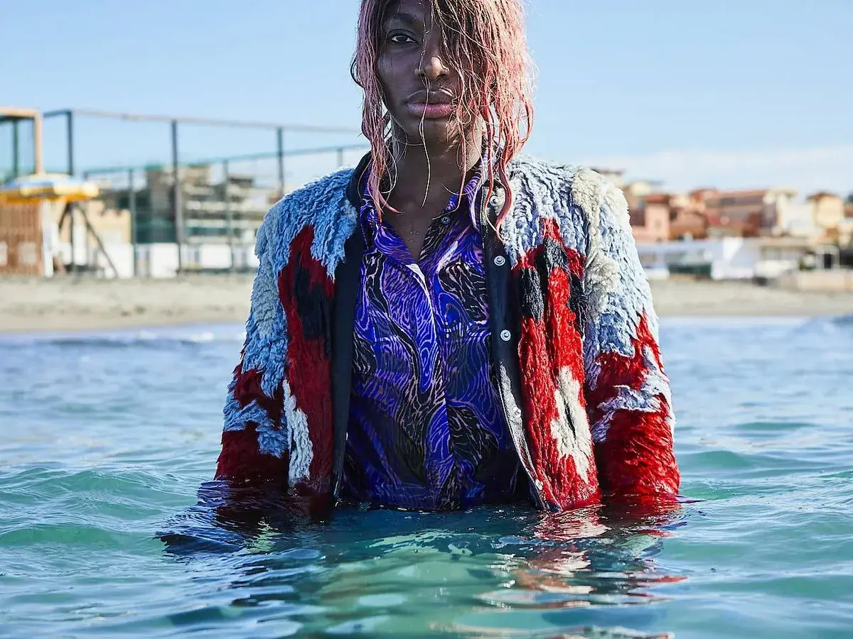 Ein Model mit pinken Haaren steht im Meer, trägt eine bunte Jacke und ein blaues Hemd. Die Wellen umspielen sie, als ob sie sagen: "I may destroy you.